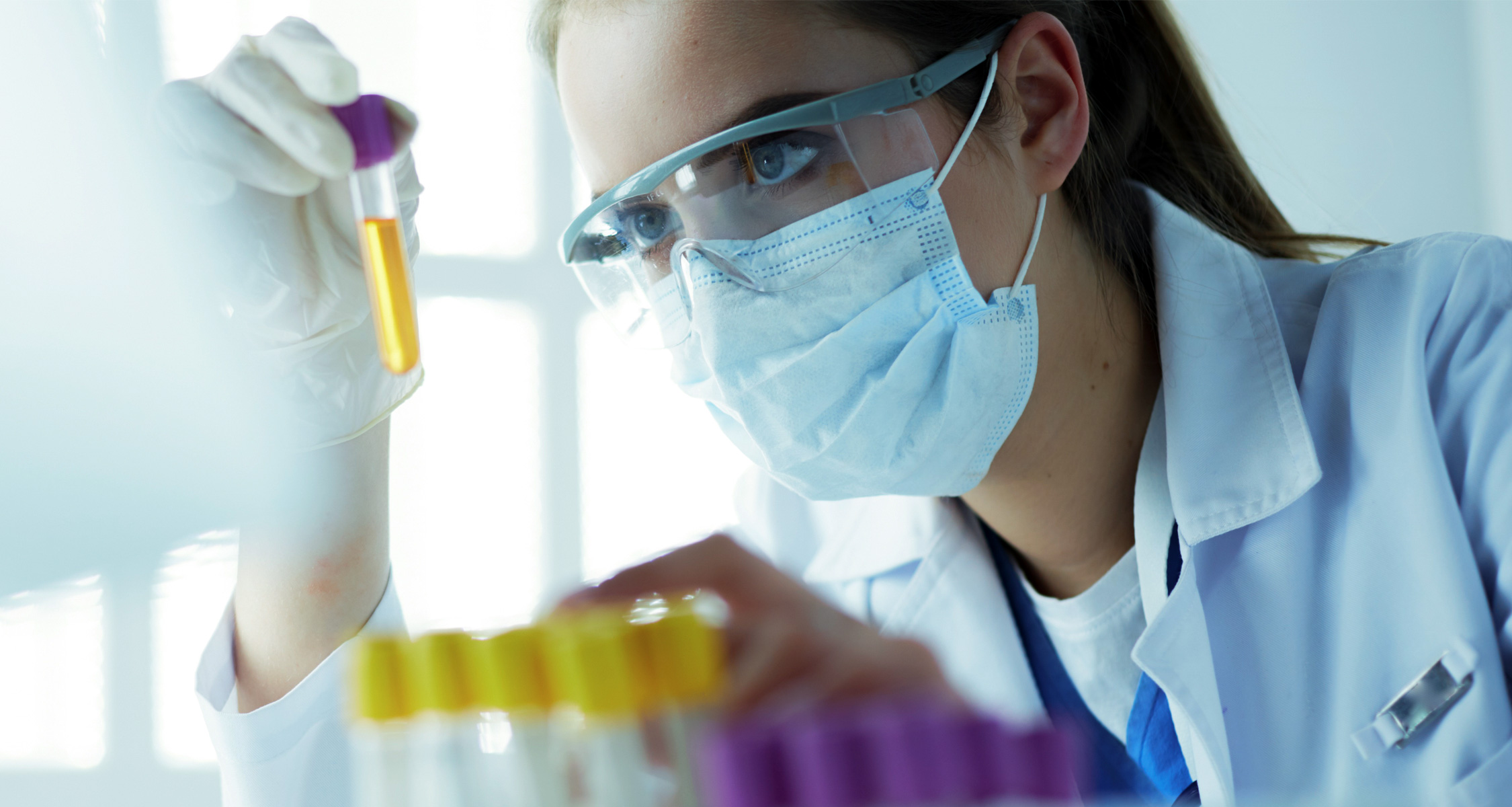 Lab technician with face mask, goggles and lab coat looks into test tube with yellow liquid.