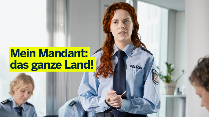 Junge Anwältin mit Polizeiuniform in Raum mit anderen Beamten. Darauf der Claim: Mein Mandant, das ganze Land!