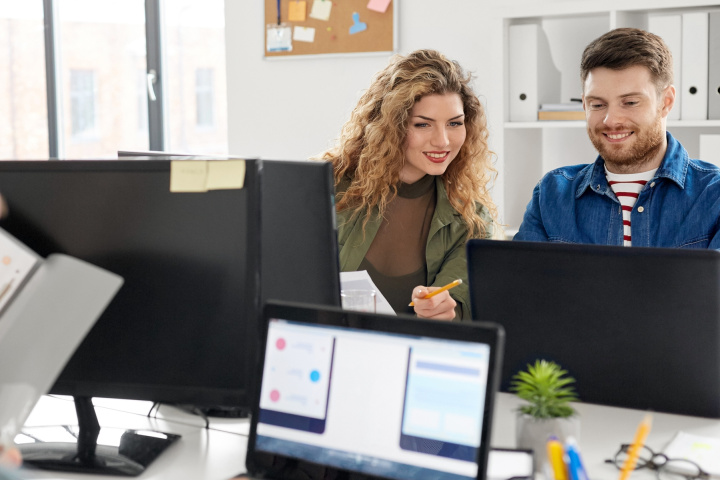 A man and a woman are looking at something together on a laptop in an office room.