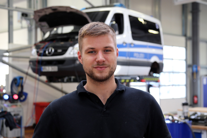 Ein junger Auszubildender steht in einer Werkstatt vor einem aufgebockten Polizeiauto.