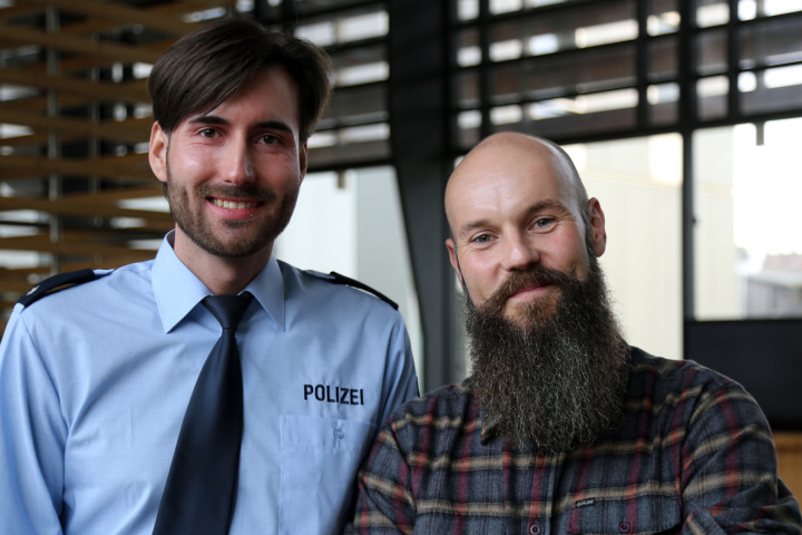 Ein Mann in Polizeiuniform und ein Mann in Zivilkleidung stehen in einem Lagerraum und lächeln freundlich.