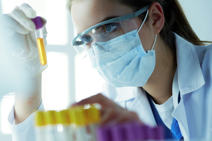 Eine Labortechnikerin mit Gesichtsmaske, Schutzbrille und Laborkittel schaut in ein Reagenzglas mit gelber Flüssigkeit.