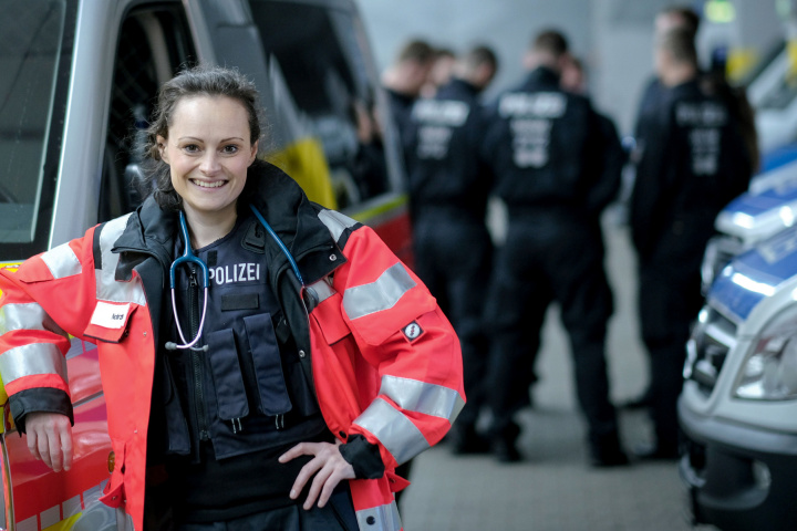 Polizeiärztin mit roter Jacke lehnt an einem Rettungsfahrzeug.