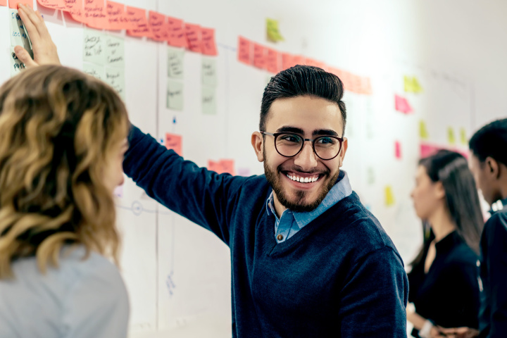 Ein Mann und eine Frau stehen vor einem Whiteboard. Der Mann zeigt darauf und dreht sich gleichzeitig weg