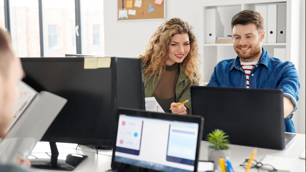 A man and a woman are looking at something together on a laptop in an office room.