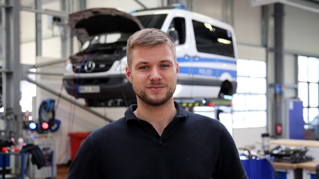 Ein junger Auszubildender steht in einer Werkstatt vor einem aufgebockten Polizeiauto.