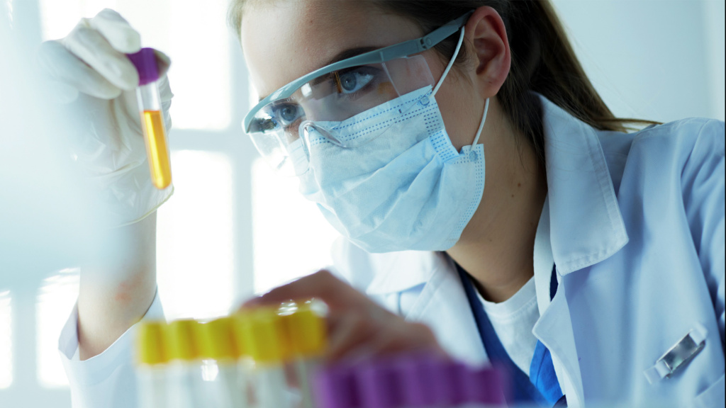 Lab technician with face mask, goggles and lab coat looks into test tube with yellow liquid.