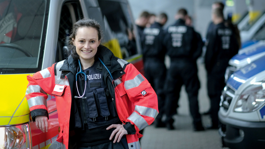 Polizeiärztin mit roter Jacke lehnt an einem Rettungsfahrzeug.