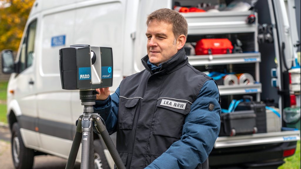 A man from the State Office of Criminal Investigation stands with a measuring device in front of a vehicle filled with technical equipment.