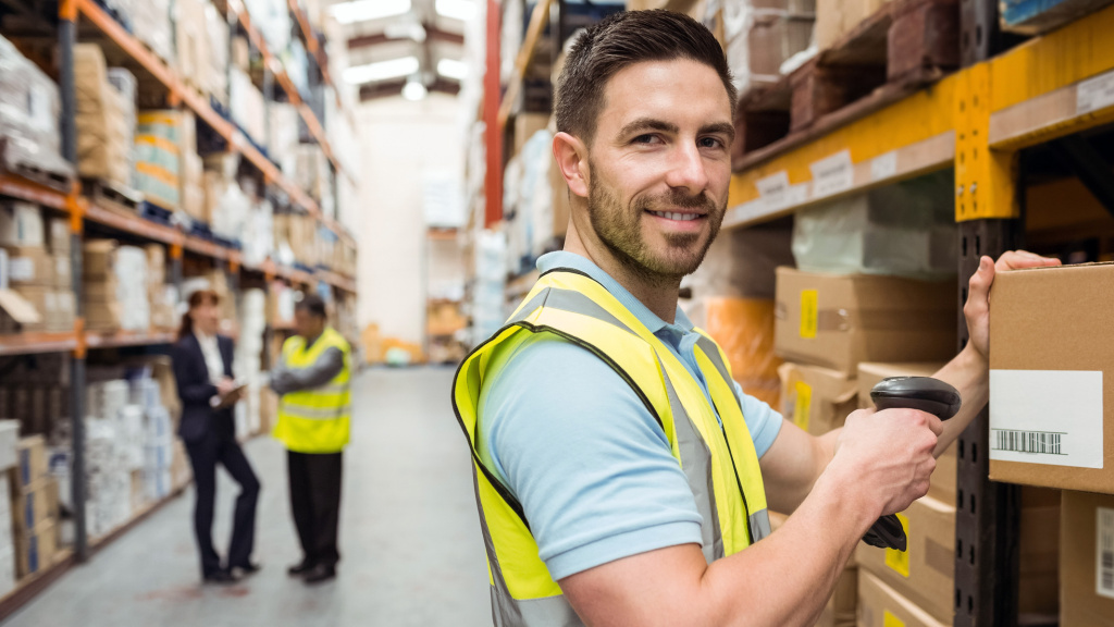 Man grasps a cardboard box on a shelf in a large warehouse. There are two other people talking in the background.