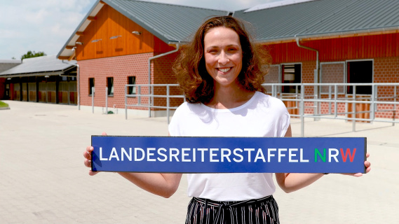 Mrs. Sobota is holing a sign "Landesreiterstaffel" in hear hand in front of a horse relay