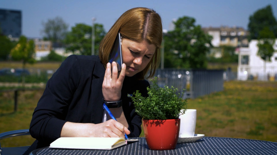 Mrs. Menke is sitting at a table on a terrace making notes in a book.